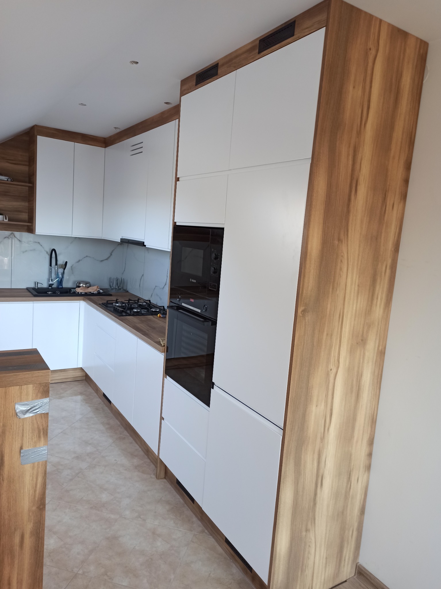 Nowoczesna kuchnia z białymi frontami i drewnianymi akcentami, zabudowa kuchenna z piekarnikiem i płytą gazową, marmurowy backsplash.