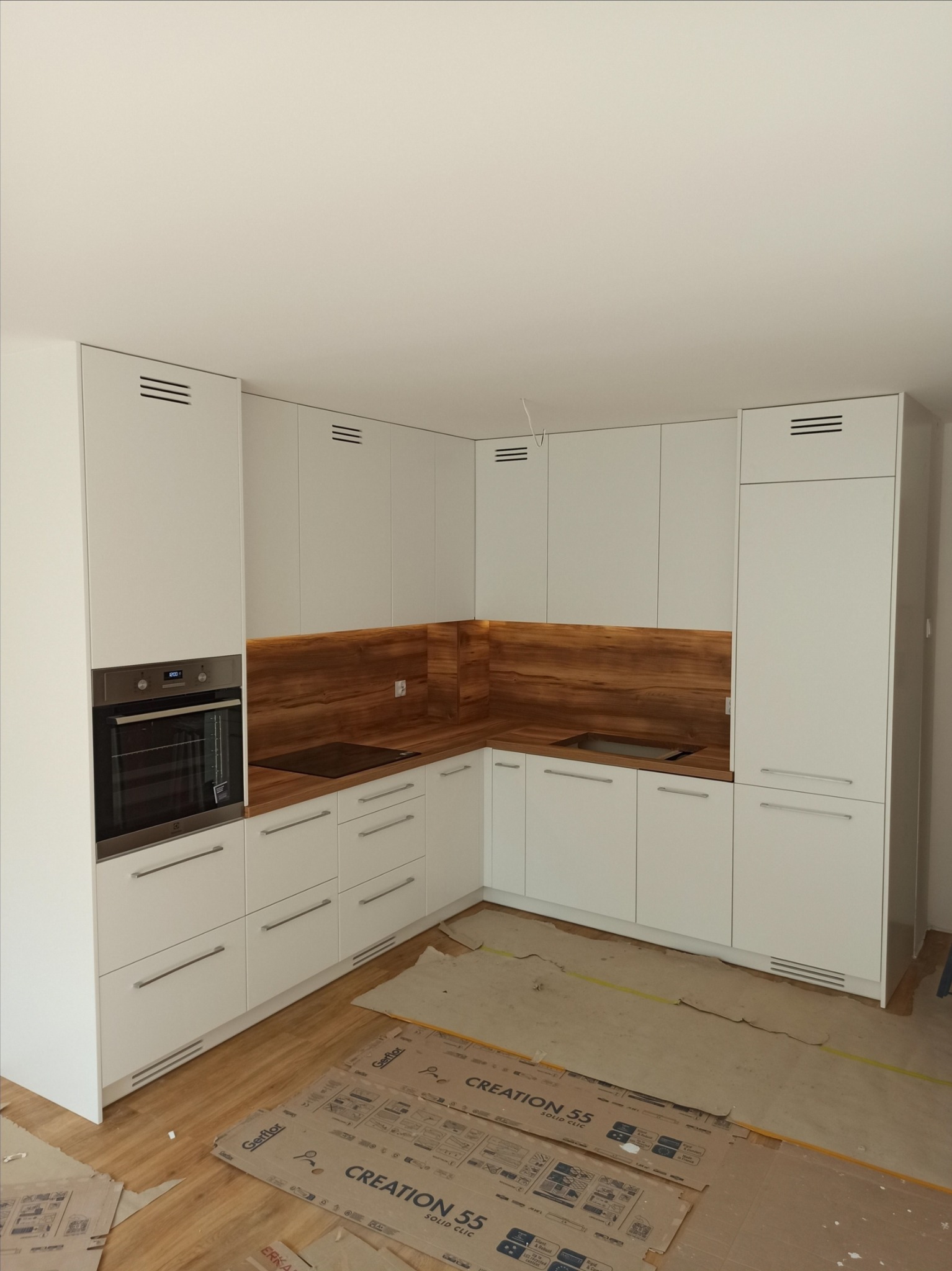 Nowoczesna, biała kuchnia z drewnianym blatem i panelem ściennym, z piekarnikiem w zabudowie i płytą indukcyjną, podłoga częściowo zabezpieczona kartonem.