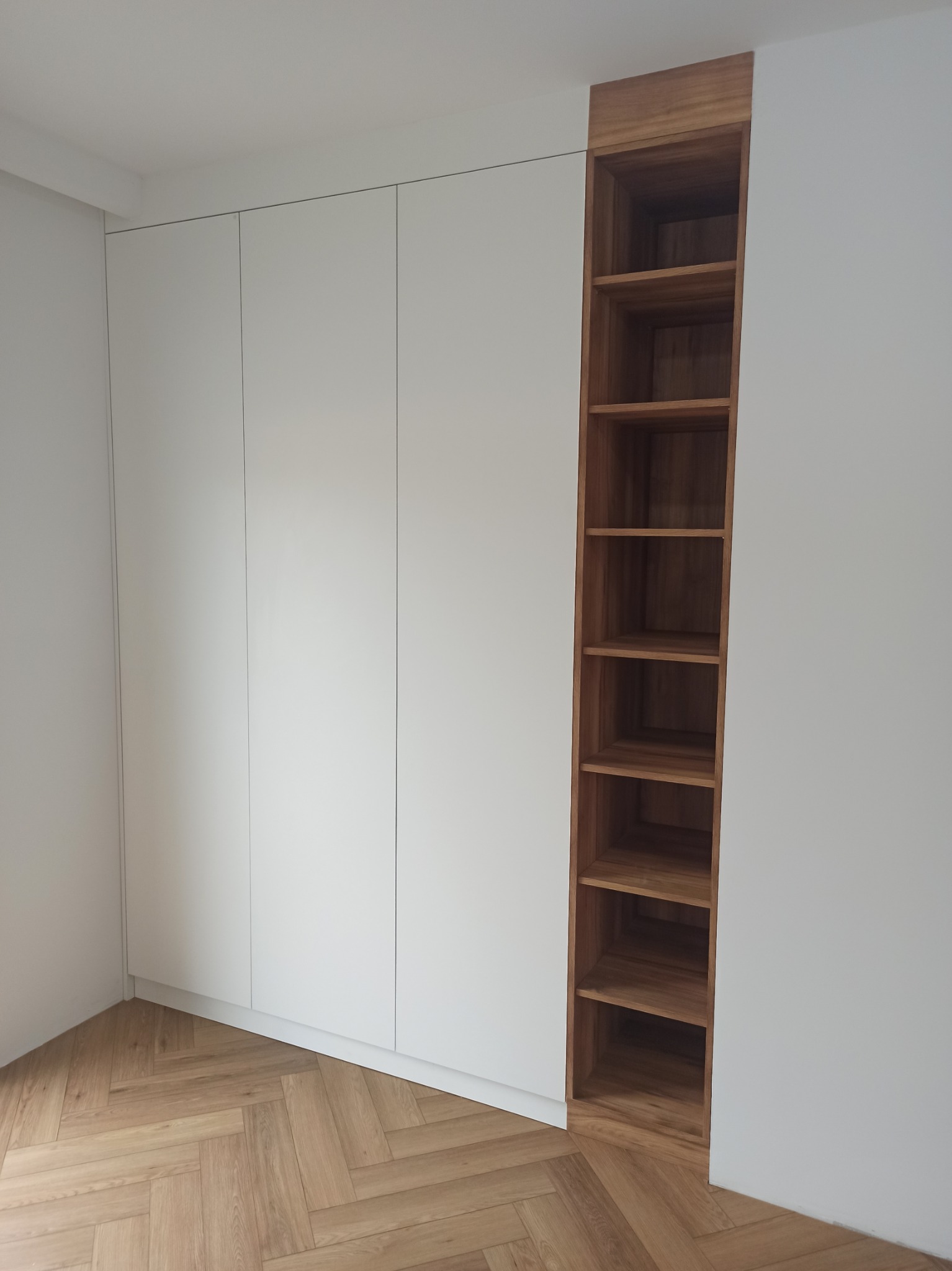 Biała zabudowa meblowa z szafkami i otwartym regałem z drewnianymi półkami, parkiet ułożony w jodełkę.