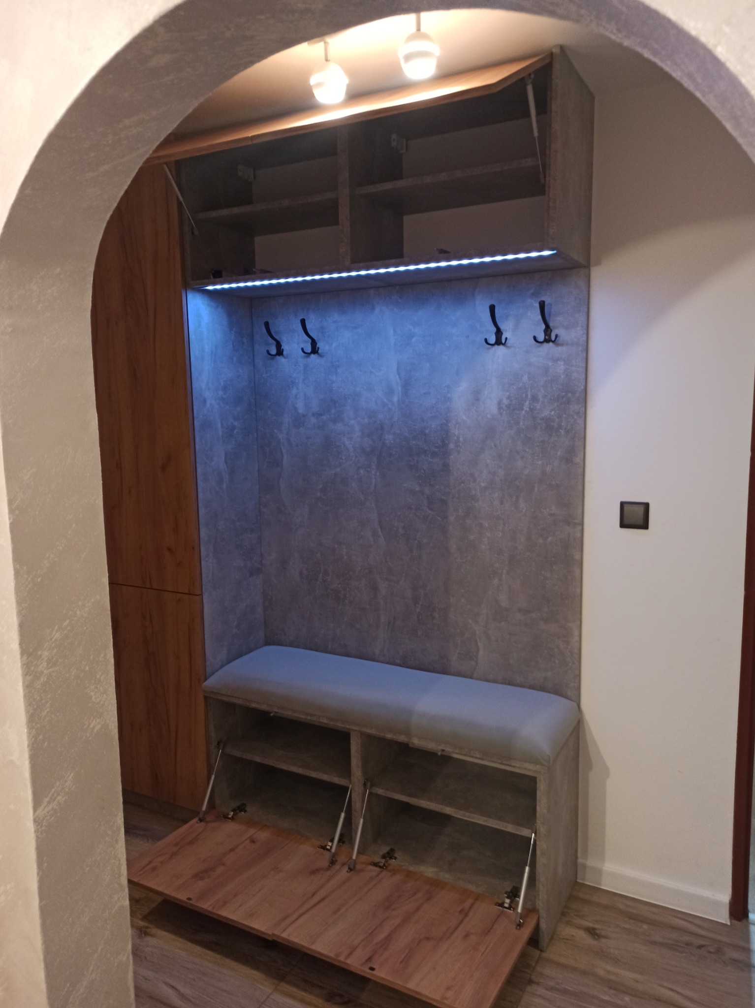 Wnęka w przedpokoju z siedziskiem, szafkami górnymi i wieszakami, oświetlona taśmą LED, widziana zza łuku.