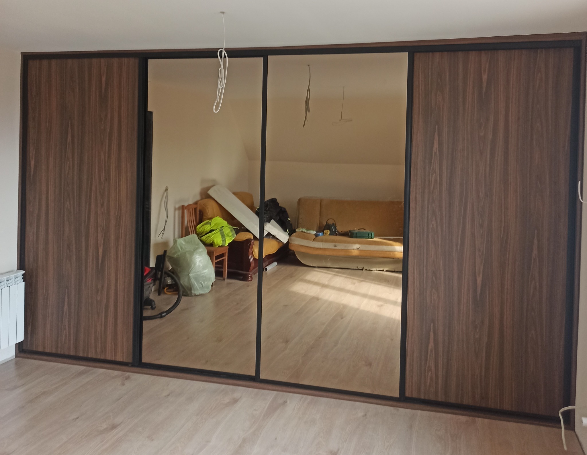 Trzydrzwiowa szafa przesuwna z lustrami i ciemnobrązowymi panelami drewnopodobnymi, odbijająca zagracony pokój z beżową sofą i workiem na podłodze.
