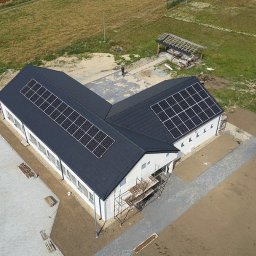 SOLARBUD - Widok z drona na budynek z panelami słonecznymi na dachu, otoczony terenem z kostką brukową i zielenią. Na ziemi widoczne rusztowania.