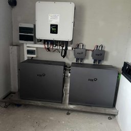 SOLARBUD - Dwa szare magazyny energii Fox ESS na metalowej platformie, z białym inwerterem Solarbud i rozdzielnicą na szarej ścianie. Widoczne kable i przewody.