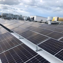 SOLARBUD - Rozległa instalacja fotowoltaiczna na dachu budynku z widocznymi elementami wentylacji i osobami w tle, pochmurne niebo.