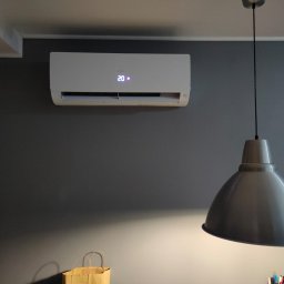 AT ADANIK SPÓŁKA Z OGRANICZONĄ ODPOWIEDZIALNOŚCIĄ - Biały klimatyzator z wyświetlaczem 20 stopni Celsjusza zamontowany na szarej ścianie, oświetlony przez metalową lampę wiszącą. Torba papierowa w dolnym rogu.