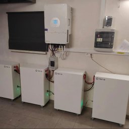 SOLARBUD - Instalacja magazynów energii Power Lab w pomieszczeniu z roletą i rozdzielnicą. Widoczne cztery białe moduły na kółkach, inwerter i okablowanie.