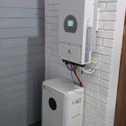 Biały inwerter fotowoltaiczny i magazyn energii Deye zamontowane na ścianie z białej cegły wewnątrz pomieszczenia. Widoczne przewody i skrzynka elektryczna.