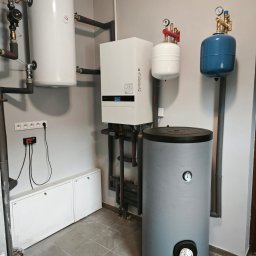 Kompletna instalacja grzewcza w kotłowni: biały piec, zbiorniki (biały, niebieski, szary), rury. Widoczne manometry i zawory. Szara ściana i płytki podłogowe.