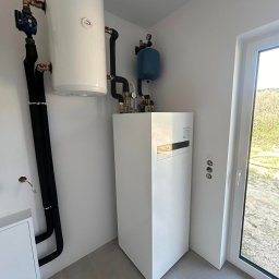 Biała pompa ciepła z zasobnikiem i instalacją w jasnym pomieszczeniu z oknem. Widoczne rury, zawory i manometry. Minimalistyczny design, szare płytki podłogowe.