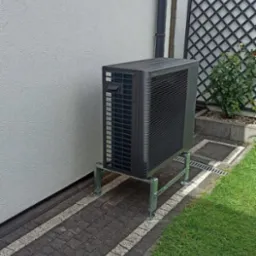 Zewnętrzna jednostka urządzenia grzewczego zamontowana na metalowej konstrukcji przy ścianie budynku, obok trawnika i ozdobnej kratki ogrodowej.