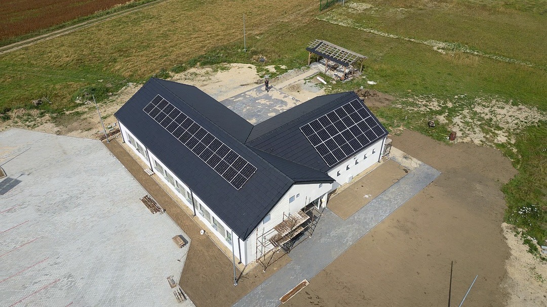 Widok z drona na budynek z panelami słonecznymi na dachu, otoczony terenem z kostką brukową i zielenią. Na ziemi widoczne rusztowania.