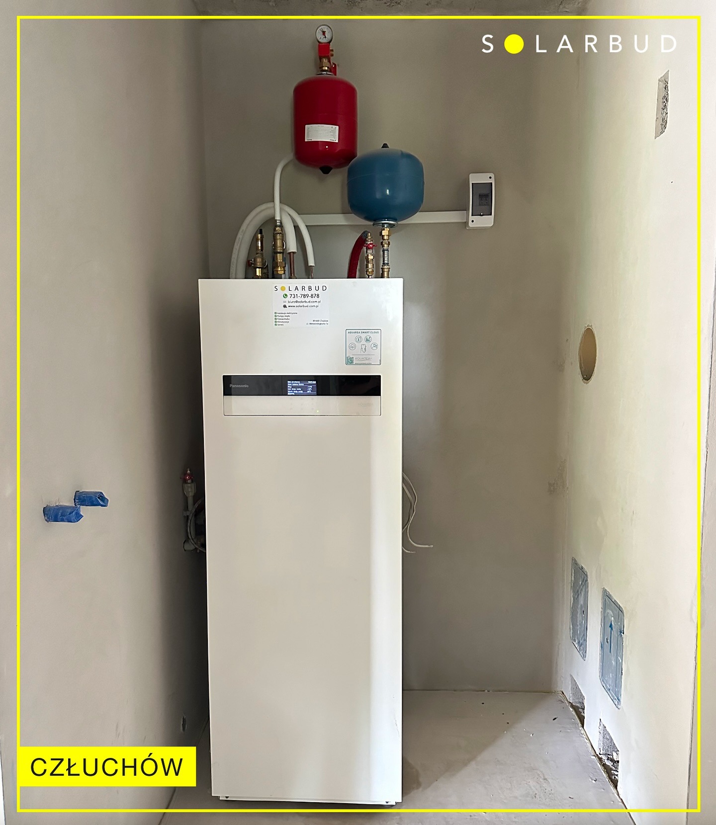 Biała pompa ciepła Panasonic z czerwonym i niebieskim zbiornikiem wyrównawczym w surowym pomieszczeniu. Widoczne instalacje i otwory w ścianie.