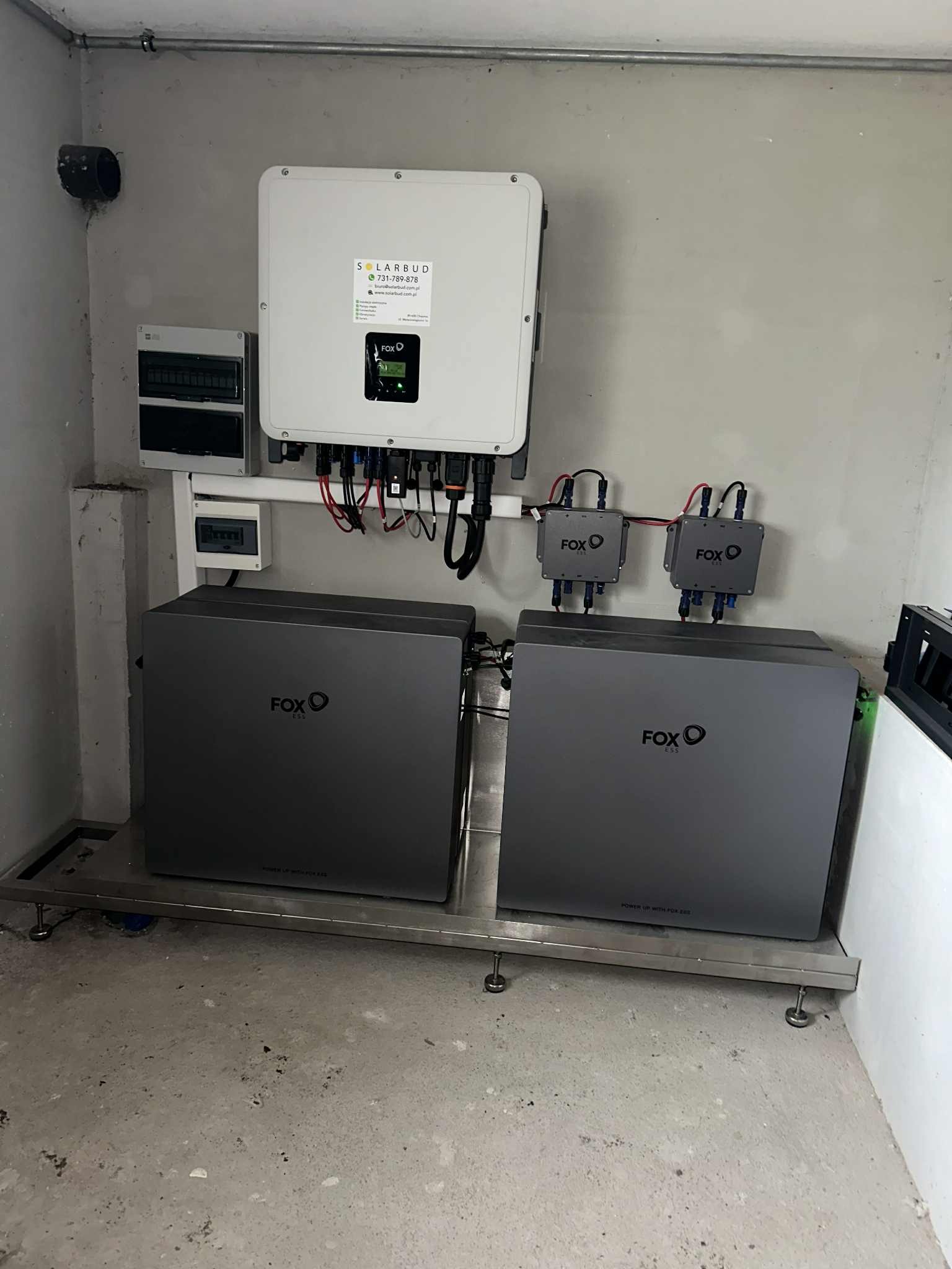 Dwa szare magazyny energii Fox ESS na metalowej platformie, z białym inwerterem Solarbud i rozdzielnicą na szarej ścianie. Widoczne kable i przewody.