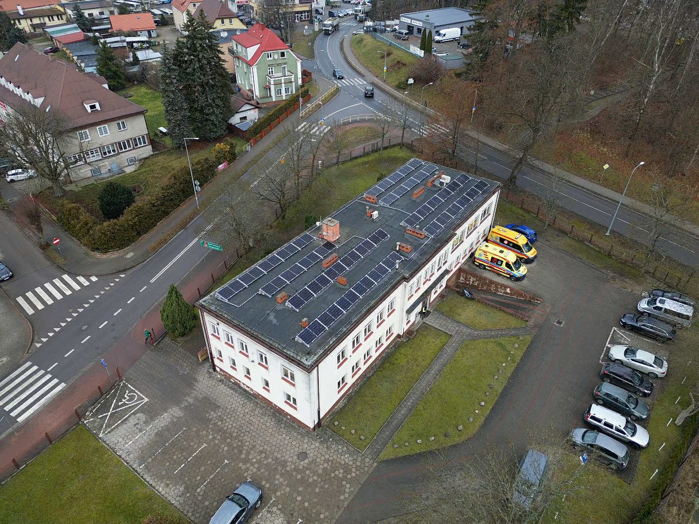 Widok z drona na budynek z panelami słonecznymi na dachu, zaparkowane ambulanse i samochody. Ujęcie z góry ukazuje otoczenie budynku: ulice, drzewa i inne budynki.