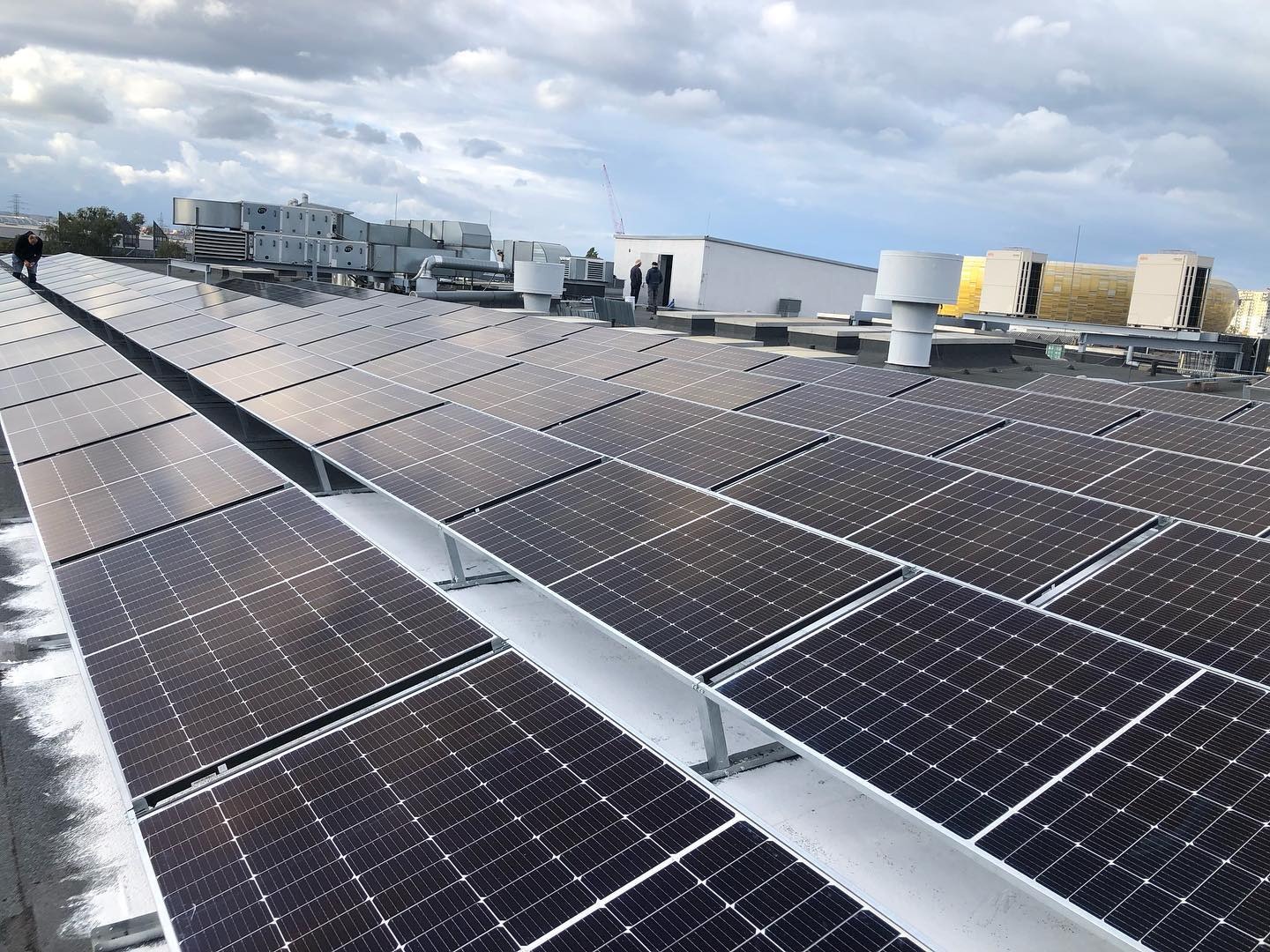 Rozległa instalacja fotowoltaiczna na dachu budynku z widocznymi elementami wentylacji i osobami w tle, pochmurne niebo.