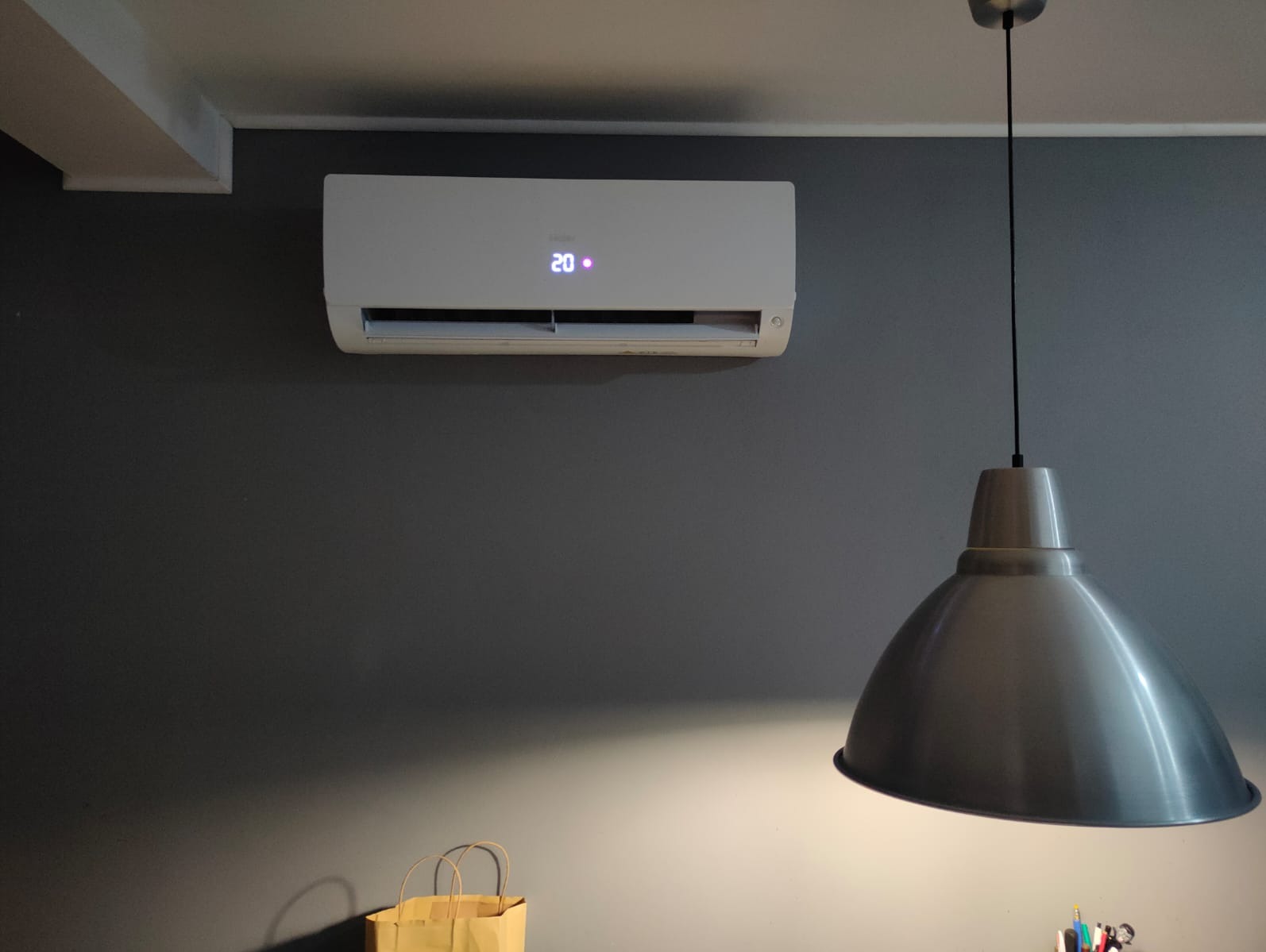 Biały klimatyzator z wyświetlaczem 20 stopni Celsjusza zamontowany na szarej ścianie, oświetlony przez metalową lampę wiszącą. Torba papierowa w dolnym rogu.
