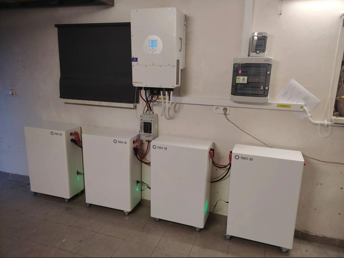 Instalacja magazynów energii Power Lab w pomieszczeniu z roletą i rozdzielnicą. Widoczne cztery białe moduły na kółkach, inwerter i okablowanie.