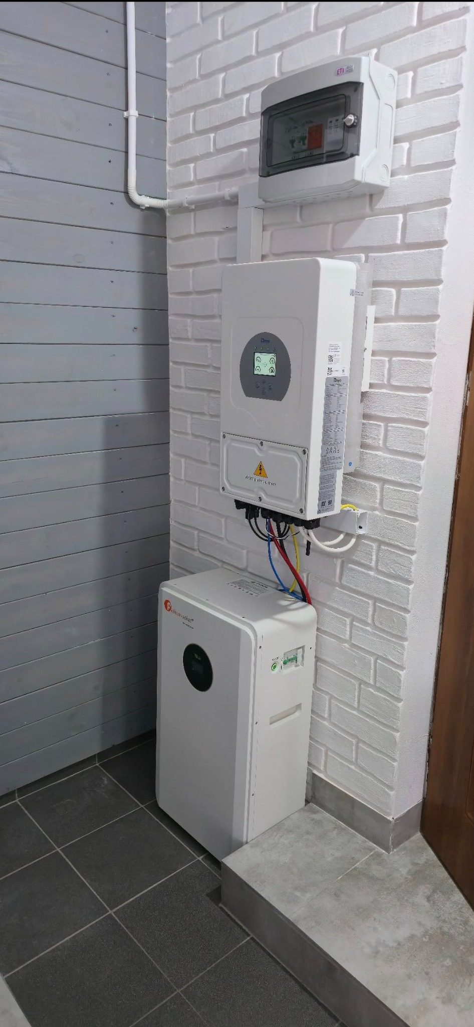 Biały inwerter fotowoltaiczny i magazyn energii Deye zamontowane na ścianie z białej cegły wewnątrz pomieszczenia. Widoczne przewody i skrzynka elektryczna.