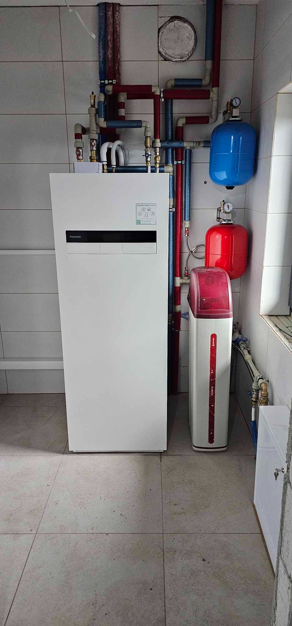 Biała pompa ciepła Panasonic Aquarea Smart Cloud z instalacją hydrauliczną, zbiornikami wyrównawczymi i zmiękczaczem wody w pomieszczeniu z szarą glazurą.