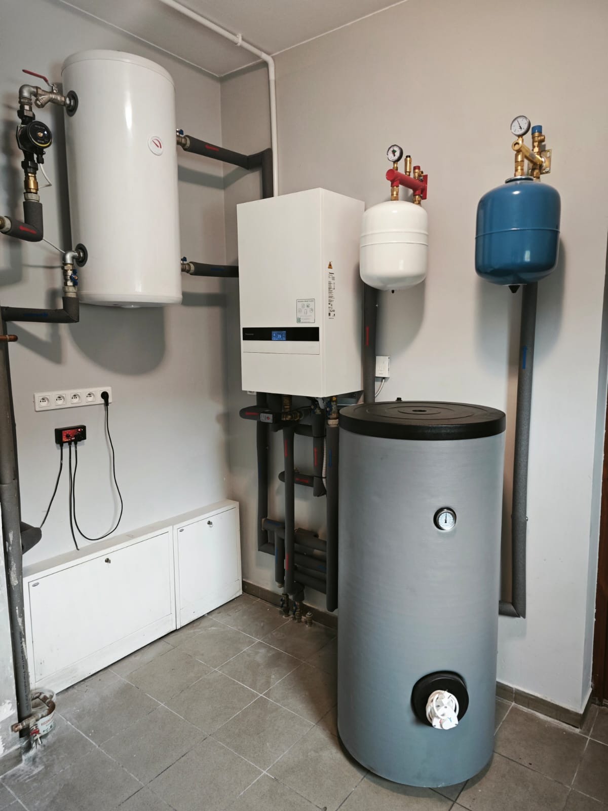 Kompletna instalacja grzewcza w kotłowni: biały piec, zbiorniki (biały, niebieski, szary), rury. Widoczne manometry i zawory. Szara ściana i płytki podłogowe.