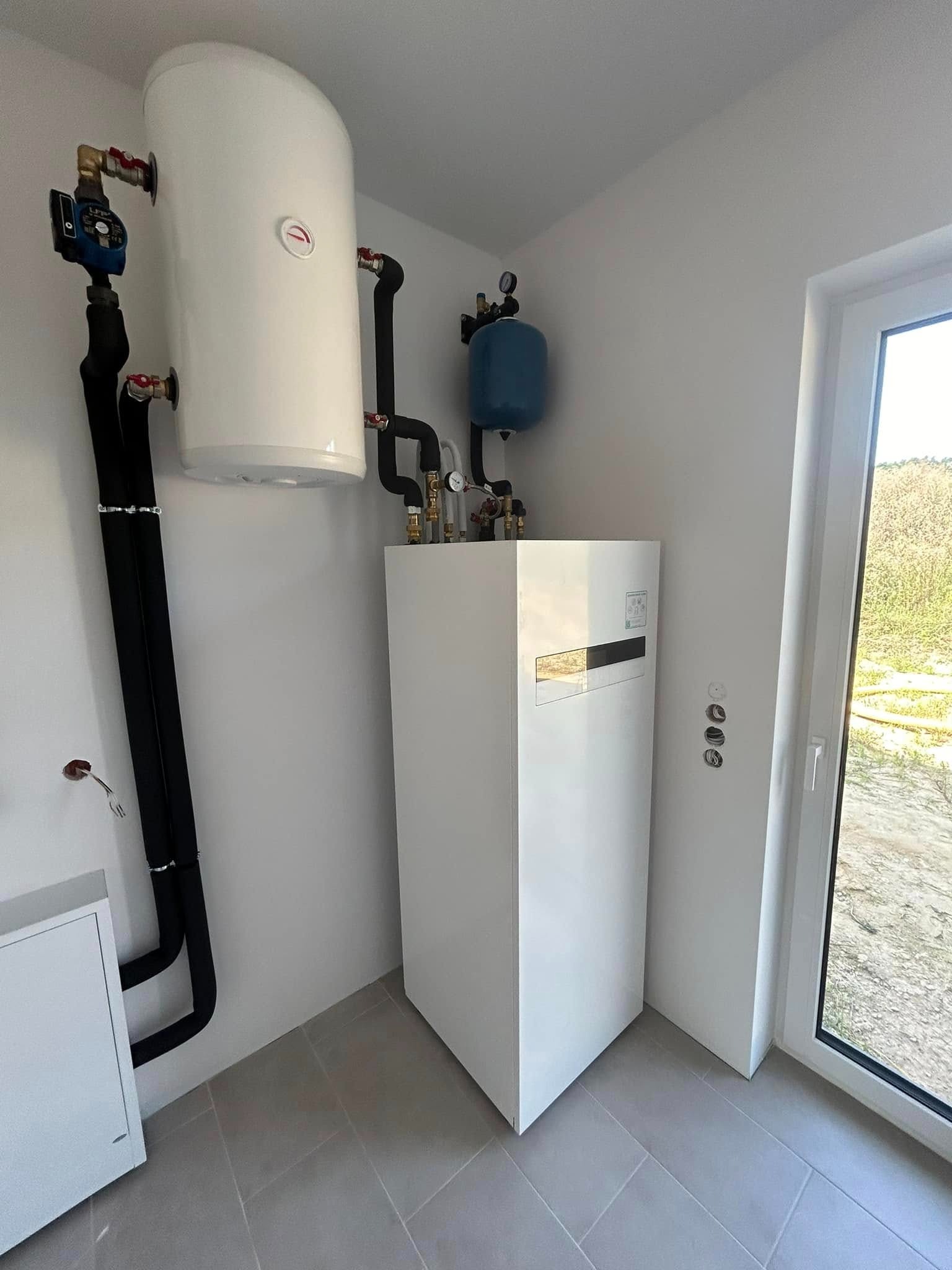 Biała pompa ciepła z zasobnikiem i instalacją w jasnym pomieszczeniu z oknem. Widoczne rury, zawory i manometry. Minimalistyczny design, szare płytki podłogowe.