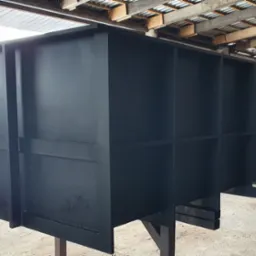 Duża, pomalowana na czarno metalowa konstrukcja z licznymi przegrodami, prawdopodobnie element bramy przemysłowej lub kontenera, na tle warsztatu.