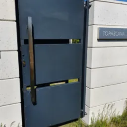Nowoczesna, grafitowa brama wjazdowa z pionowym, prostokątnym uchwytem ze stali nierdzewnej, zamontowana na betonowych słupkach ogrodzeniowych z widoczną tabliczką z numerem domu.