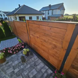 Nowoczesne ogrodzenie z drewnianych paneli w ciepłym odcieniu brązu, z czarnymi metalowymi słupkami, otoczone kwiatami i kostką brukową, na tle zabudowy jednorodzinnej.