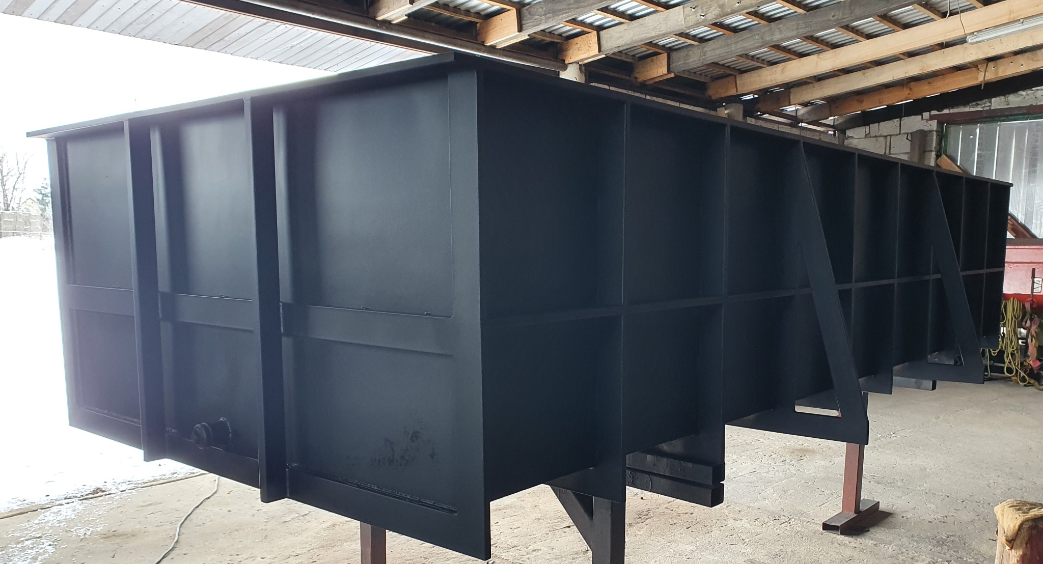Duża, pomalowana na czarno metalowa konstrukcja z licznymi przegrodami, prawdopodobnie element bramy przemysłowej lub kontenera, na tle warsztatu.