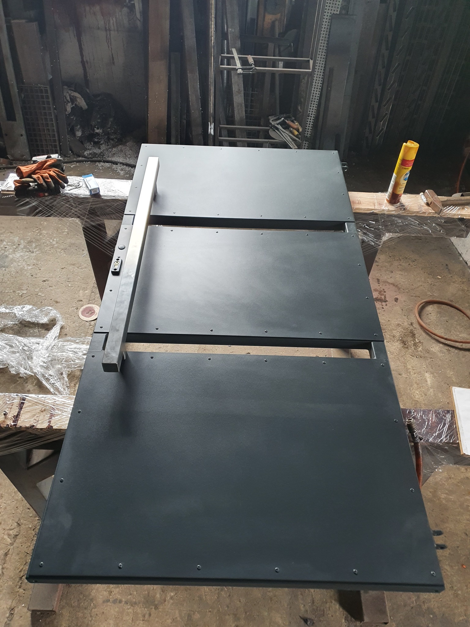 Surowa, czarna brama wjazdowa z metalowymi panelami i prostokątnym uchwytem, widoczna w warsztacie podczas montażu zamka.