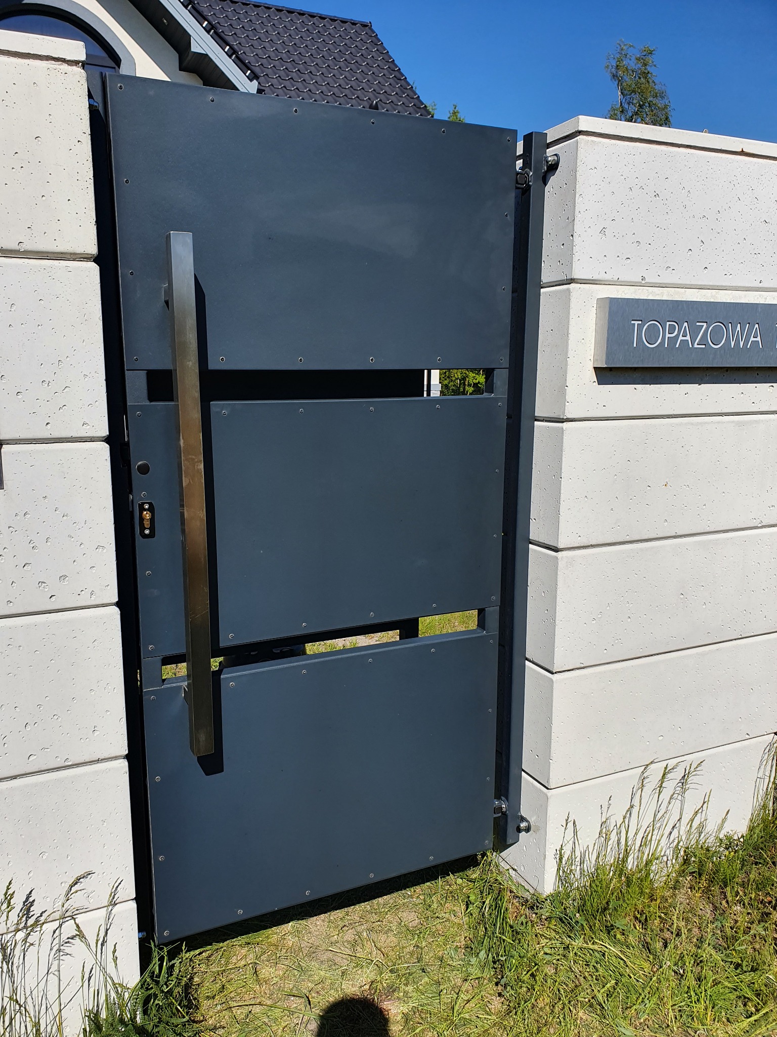 Nowoczesna, grafitowa brama wjazdowa z pionowym, prostokątnym uchwytem ze stali nierdzewnej, zamontowana na betonowych słupkach ogrodzeniowych z widoczną tabliczką z numerem domu.