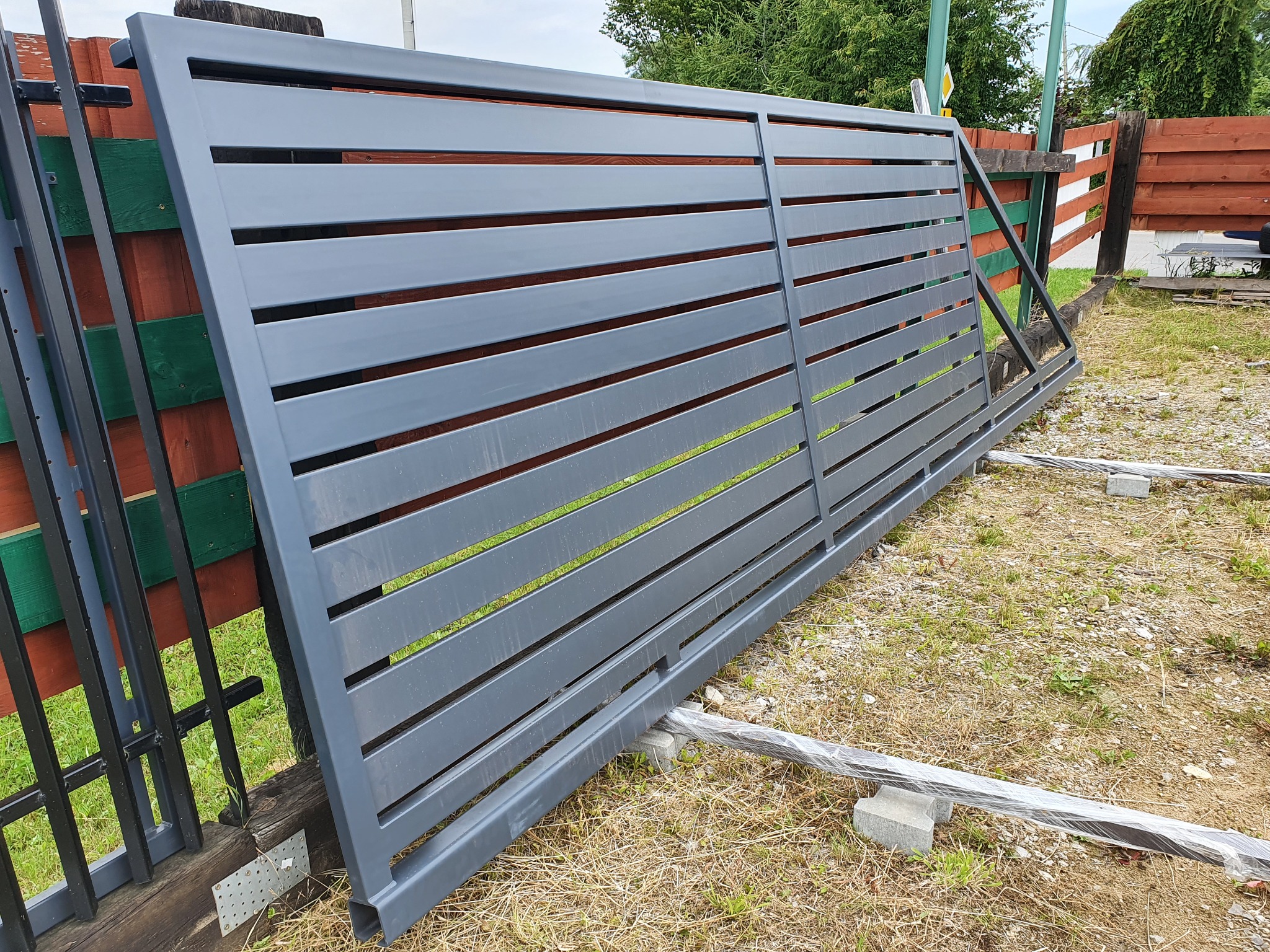 Przygotowana do montażu, pozioma, przesuwna brama wjazdowa z metalowymi panelami w kolorze szarym, oparta na betonowych bloczkach w Cycowie, widoczna na tle drewnianego ogrodzenia.