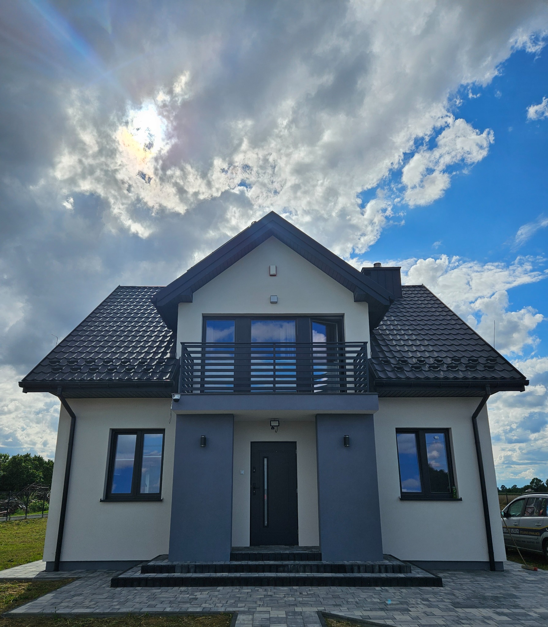 Nowoczesny, dwukondygnacyjny dom z ciemną dachówką i szarą elewacją, balkonem z metalową balustradą i brukowanym podjazdem, sfotografowany w słoneczny dzień z widocznymi chmurami odbijającymi się...