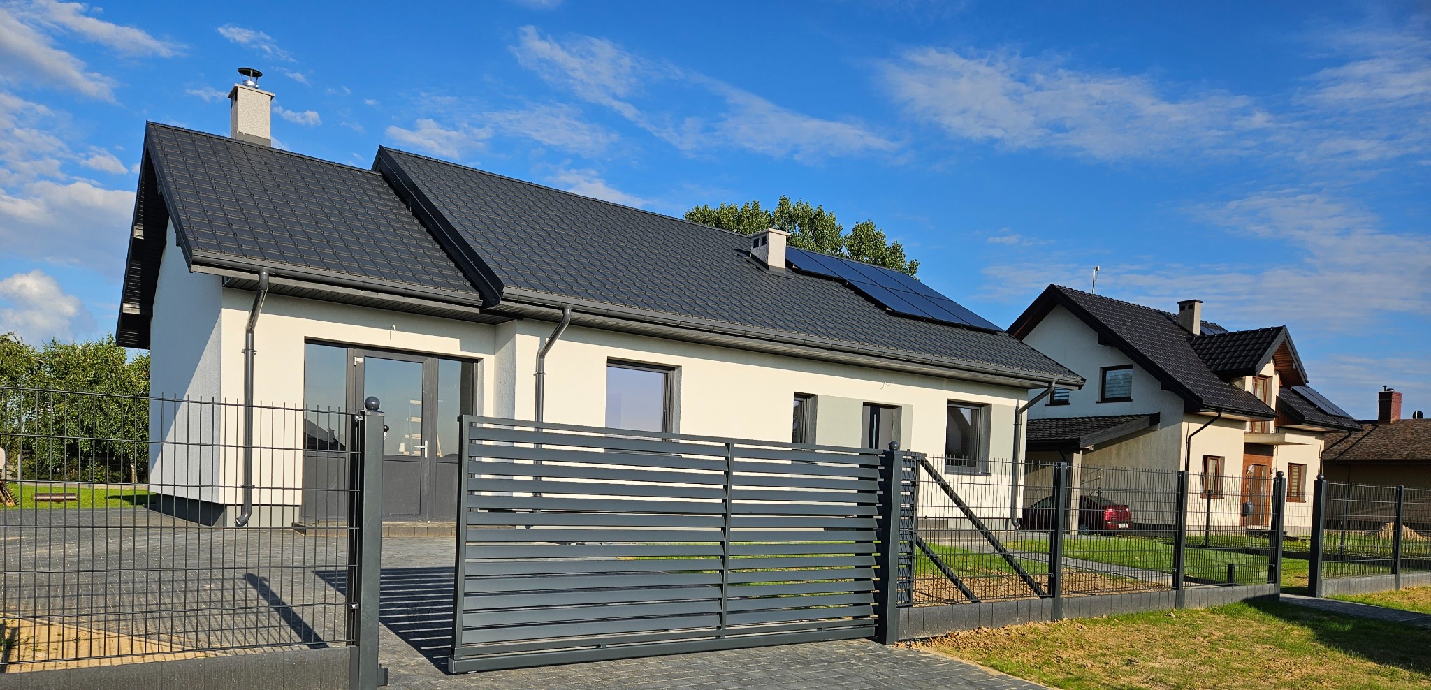 Nowoczesna posesja z szarym, poziomym ogrodzeniem panelowym i przesuwną bramą wjazdową, na tle domu z panelami słonecznymi w słoneczny dzień.
