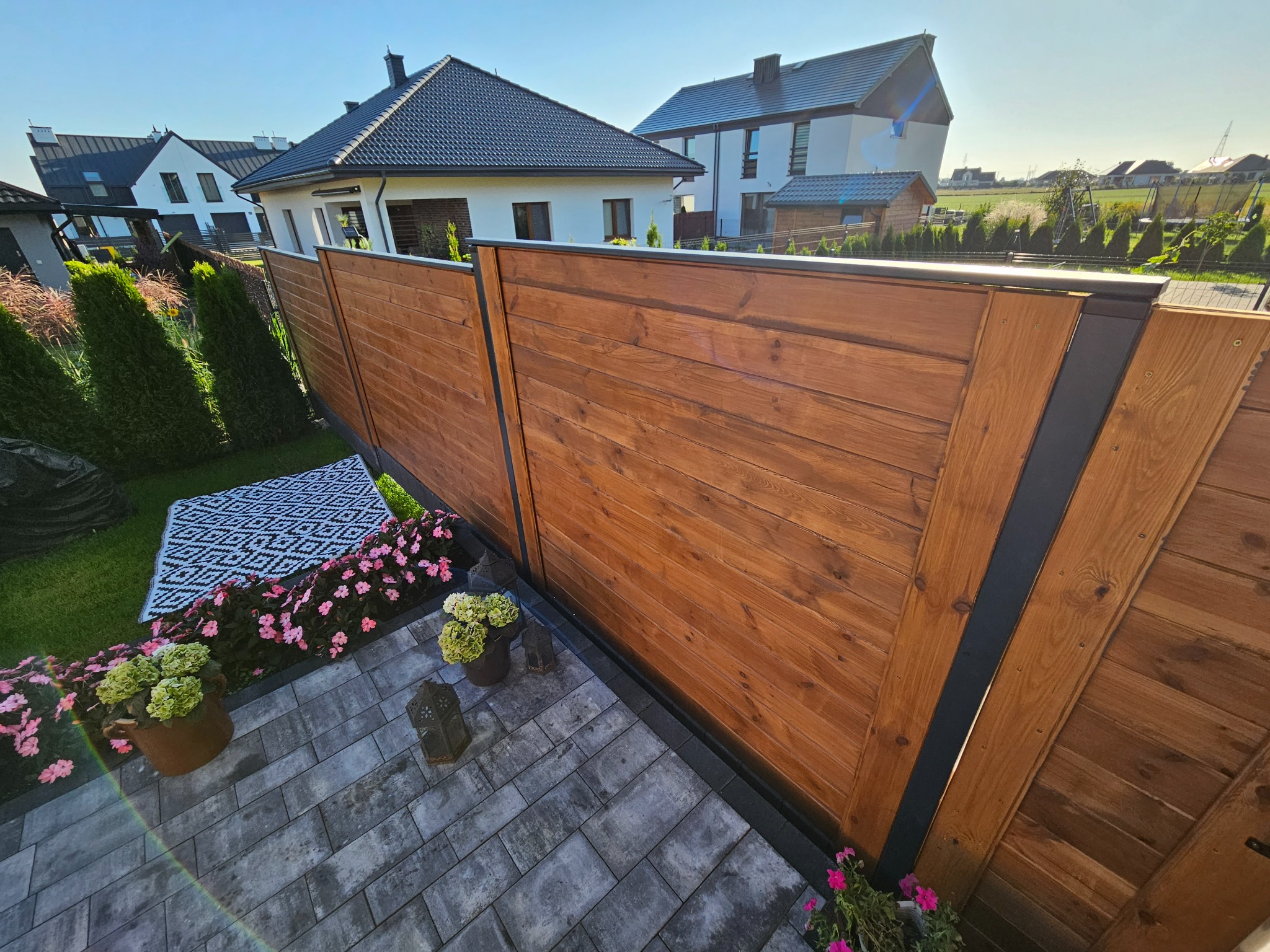 Nowoczesne ogrodzenie z drewnianych paneli w ciepłym odcieniu brązu, z czarnymi metalowymi słupkami, otoczone kwiatami i kostką brukową, na tle zabudowy jednorodzinnej.