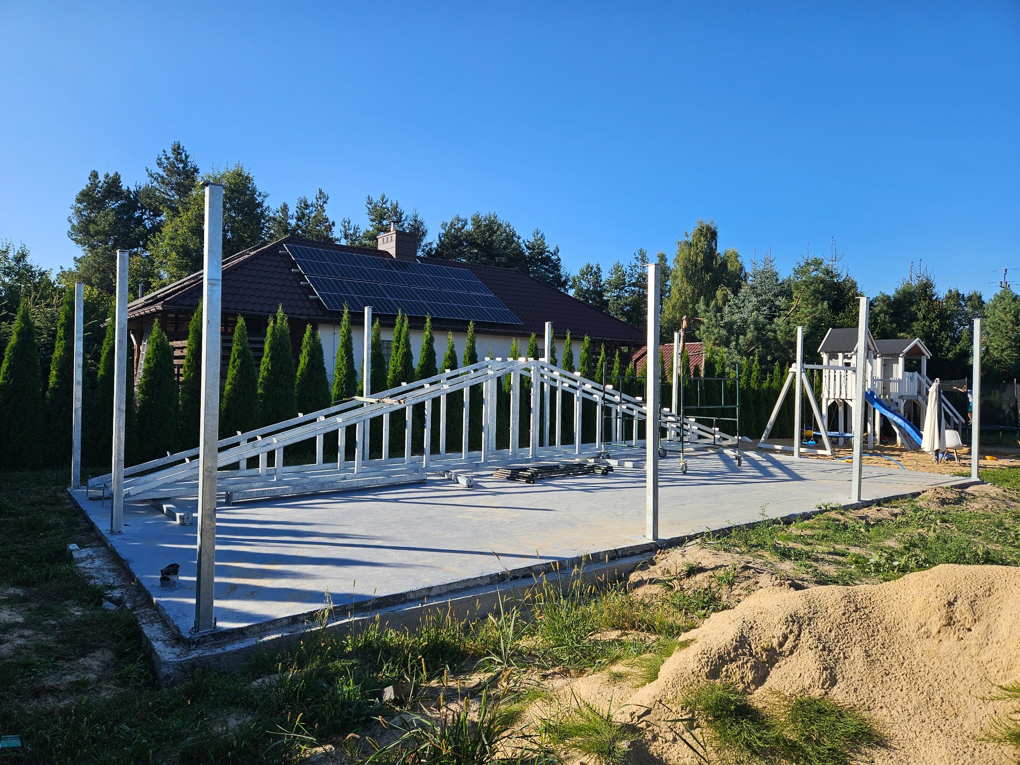 Konstrukcja metalowa z profili w trakcie montażu na wylewce betonowej, widoczna na tle domu z panelami słonecznymi i placu zabaw.