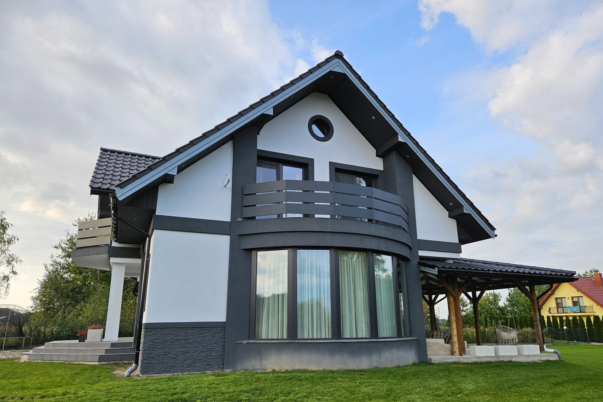 Nowoczesny dom z balkonem z metalową balustradą, czarno-biała elewacja, widok z trawnika, zadaszony taras z drewnianymi słupami w tle.