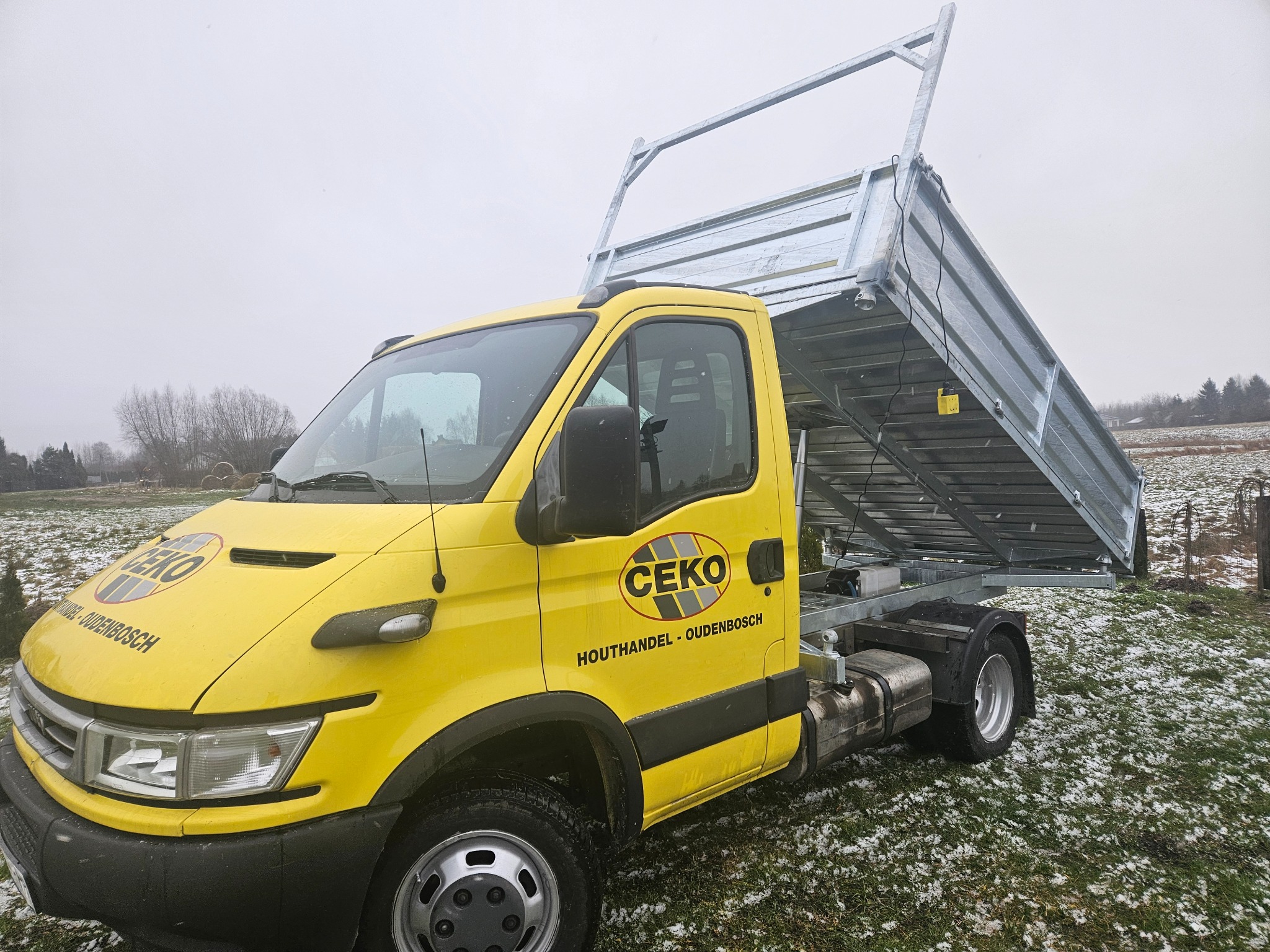 Żółty samochód dostawczy z podniesioną metalową platformą załadunkową na tle zaśnieżonej trawy i pochmurnego nieba.
