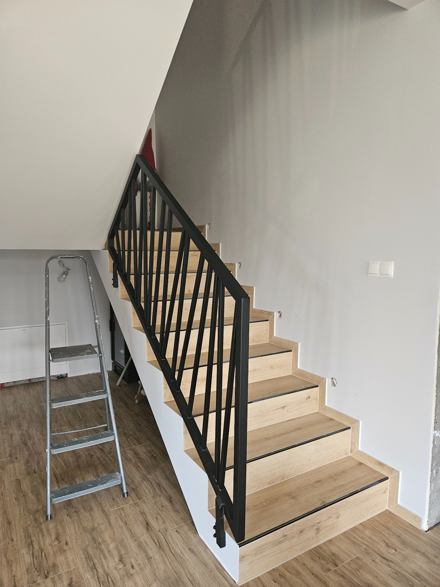 Drewniane schody z czarną, metalową balustradą o geometrycznym wzorze, widziane z dołu, z aluminiową drabiną opartą o ścianę po lewej stronie.