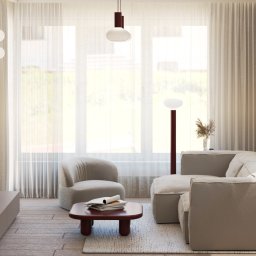 Adelina Interiors - Jasne wnętrze salonu z beżową sofą narożną, fotel z barankiem, stolik kawowy z magazynami i telewizor na ścianie z drewnianym panelem. Duże okna z delikatnymi zasłonami.