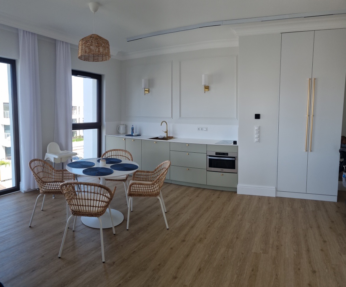 Jasna kuchnia z okrągłym stołem i wiklinowymi krzesłami, minimalistyczne szafki kuchenne w odcieniach szarości, drewniana podłoga, pleciona lampa nad stołem.