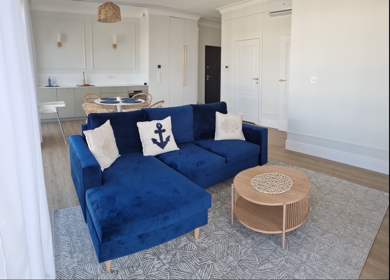 Niebieski narożnik z poduszkami (kotwica i muszla) w jasnym salonie z okrągłym stolikiem kawowym i dywanem w geometryczne wzory, widok na kuchnię z jadalnią.