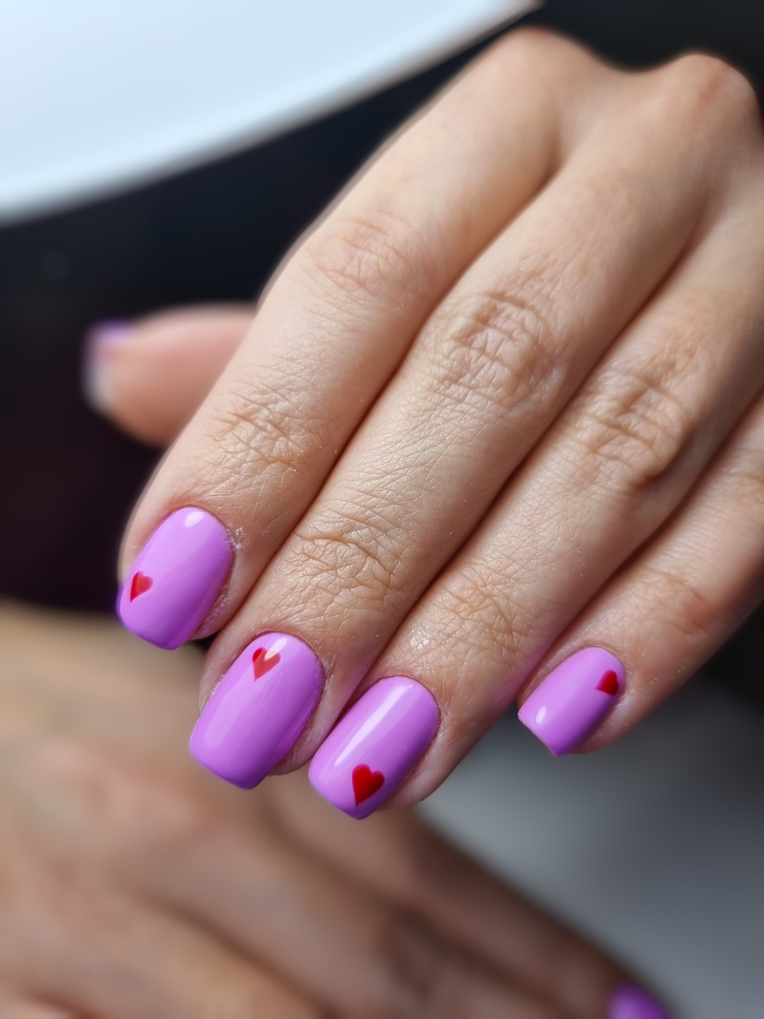 Dłoń z pomalowanymi na liliowo paznokciami w kształcie kwadratu, ozdobionymi małymi czerwonymi serduszkami, widoczne delikatne zmarszczki na skórze.