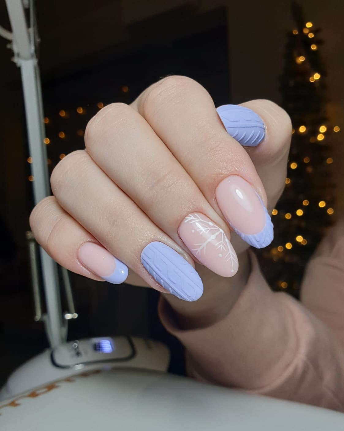 Dłoń z zimowym zdobieniem paznokci: pastelowy róż i lila french, wypukły wzór sweterka na niektórych paznokciach, na jednym biały śnieżynka, w tle lampa UV i choinka z delikatnymi światełkami.