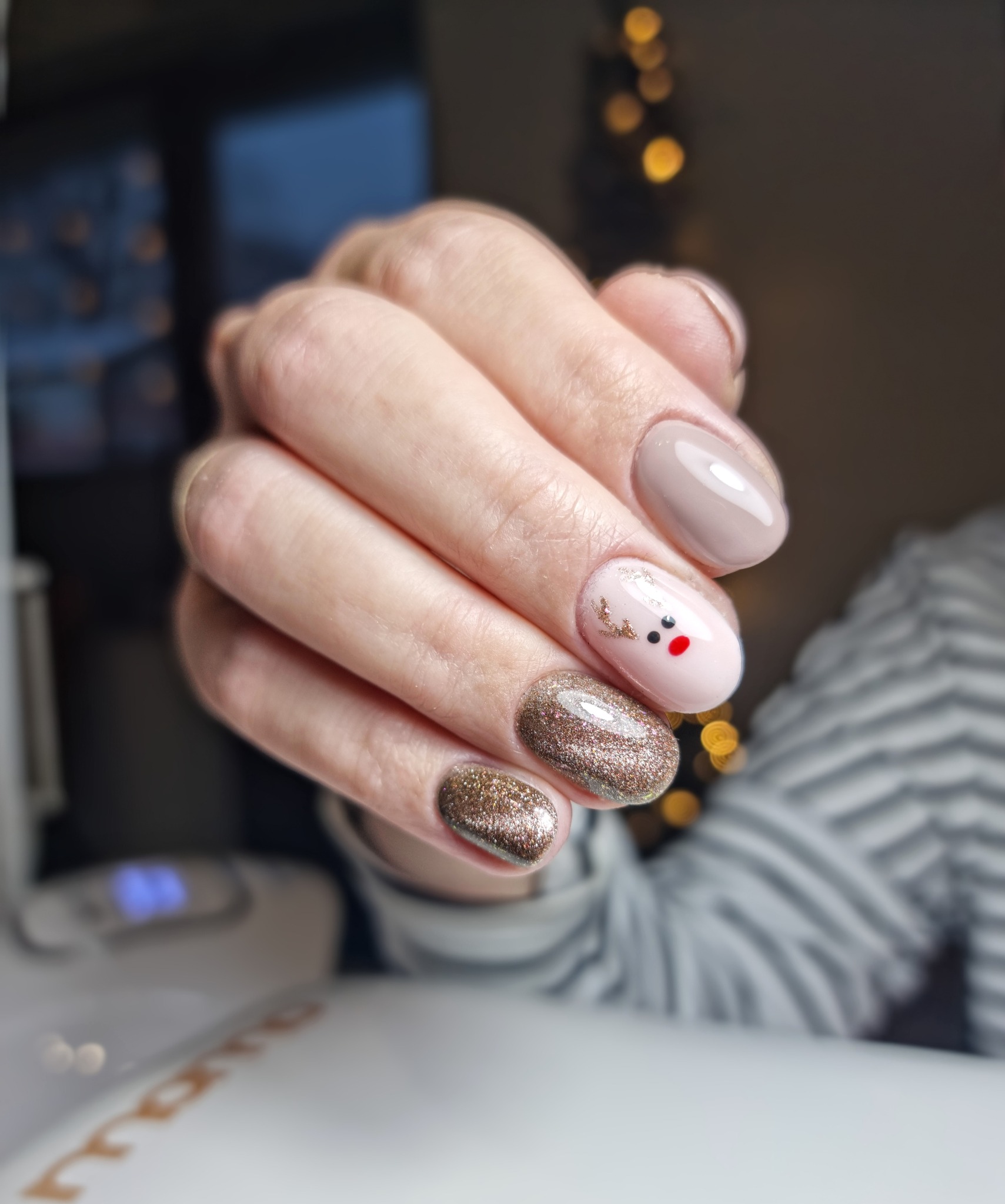 Dłoń z manicure hybrydowym: beżowy, brokatowy złoty i jasnoróżowy z motywem renifera, wykonany na tle rozmytych światełek choinkowych.