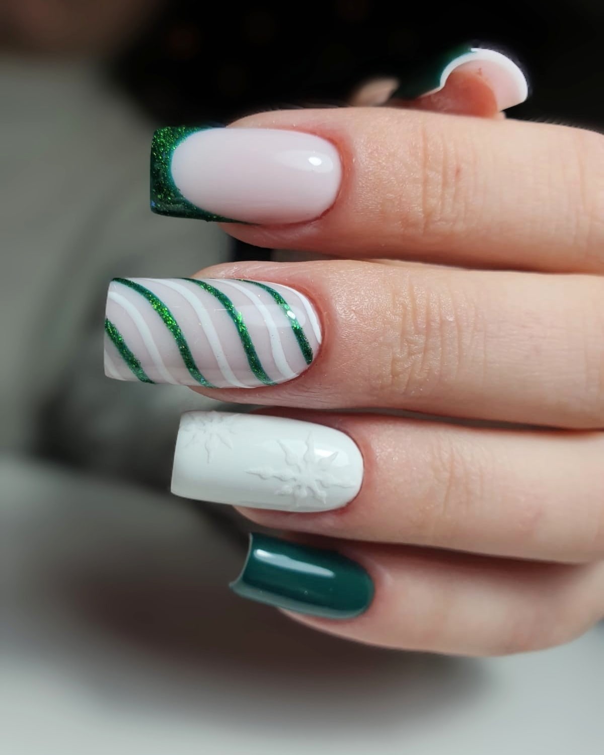Zbliżenie na dłoń z pomalowanymi paznokciami w świąteczne wzory: brokatowy, zielony french manicure, białe i zielone paski oraz biały paznokieć z wytłoczonym wzorem śnieżynki.