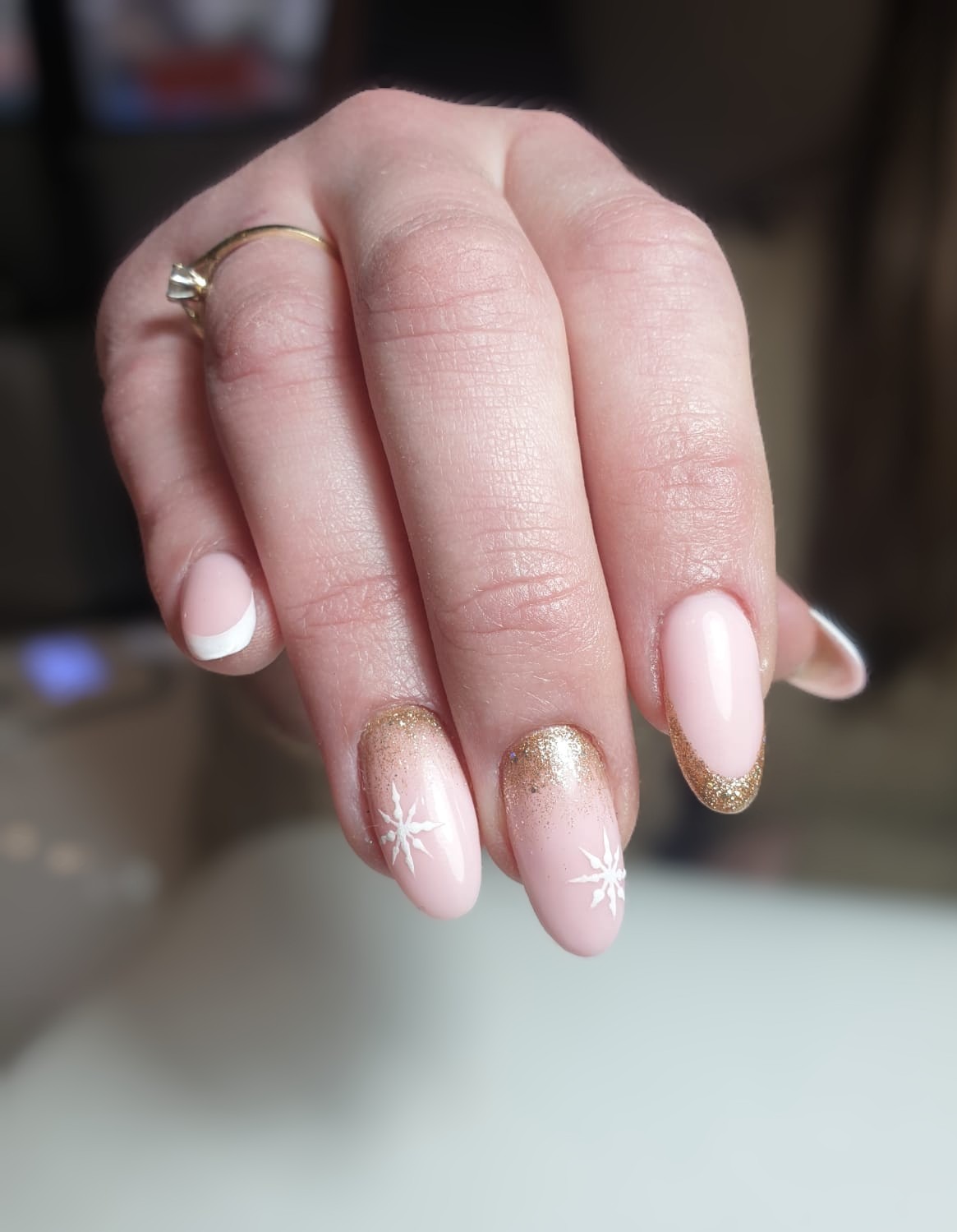 Dłoń z delikatnym, różowym manicure hybrydowym. Dwa paznokcie zdobione białymi gwiazdkami i złotym brokatem, jeden z białym francuskim, a jeden ze złotym francuskim. Kobieta ma na palcu złoty...