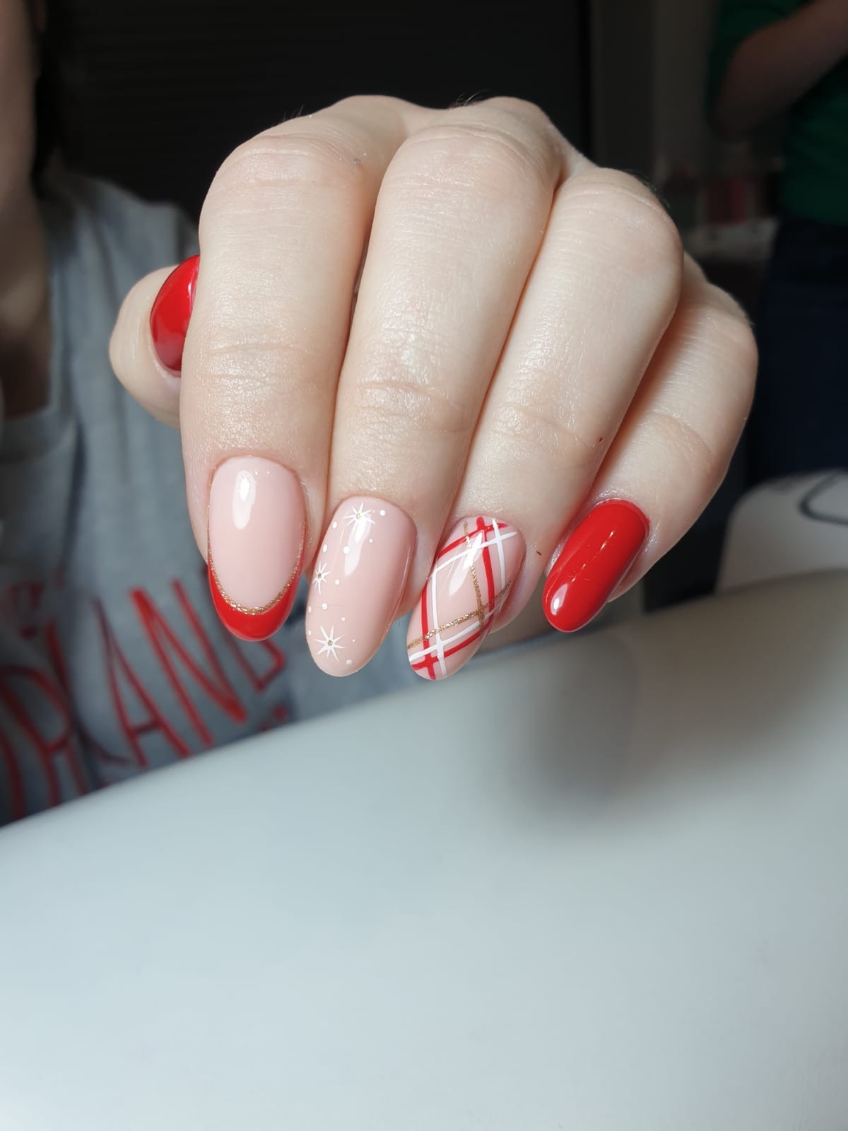 Dłoń z manicure hybrydowym: czerwone, błyszczące paznokcie oraz paznokcie w kolorze jasnego różu z motywem kratki w czerwieni i bieli ze złotymi akcentami oraz białymi gwiazdkami, na jasnym tle.