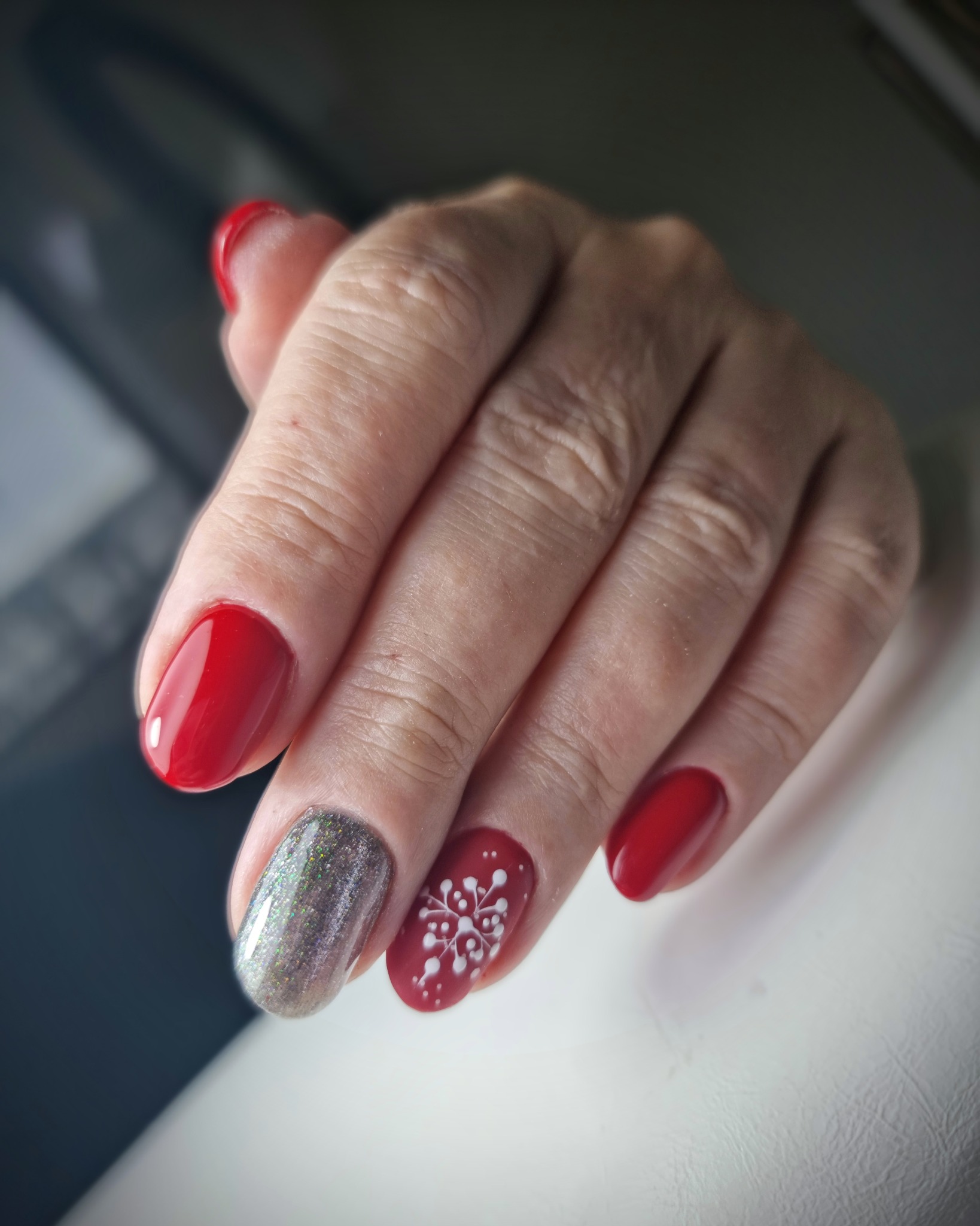 Dłoń z czerwonymi paznokciami, jeden srebrny brokatowy i jeden z białym wzorem śnieżynki na czerwonym tle.