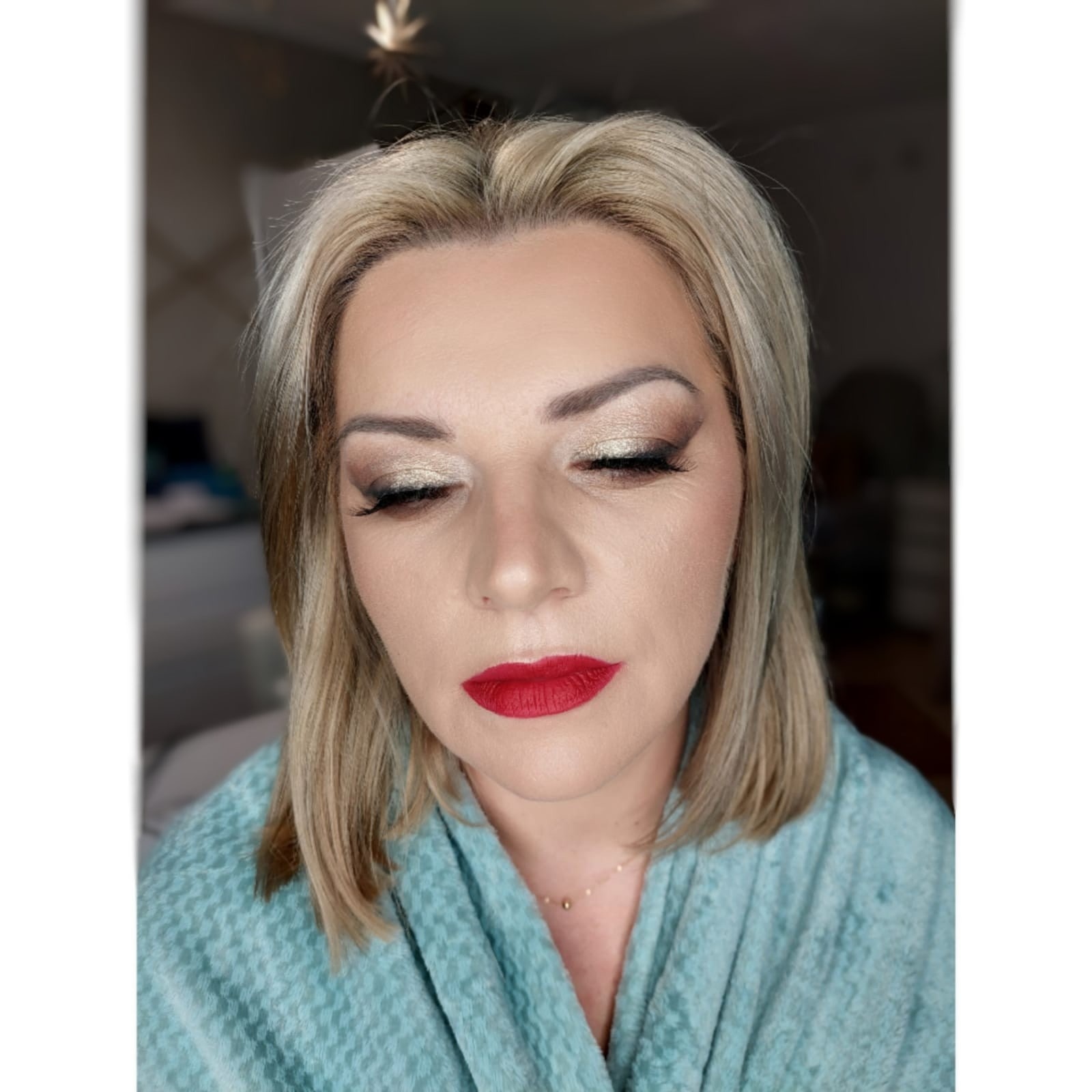 Portret kobiety z zamkniętymi oczami, w szlafroku, z wykonanym makijażem: złoty cień na powiekach, mocno wytuszowane rzęsy i intensywnie czerwona szminka na ustach.